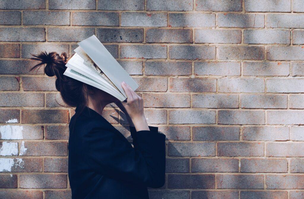 female student stood in front of a brick wall facing the side, with an academic book held up in front of her head so it's covering her face