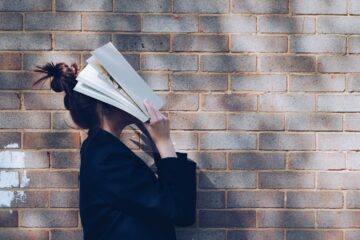 female student stood in front of a brick wall facing the side, with an academic book held up in front of her head so it's covering her face