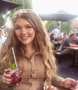 lucy dawson, who discusses encephalitis and employment, sat at an outdoor table holding a cocktail, smiling with her long hair down