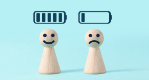 Two wooden figures of people stood next to each other. One is smiling with a full battery symbol over their head, the other is frowning with a low battery signal over their head, to represent energy limiting conditions.